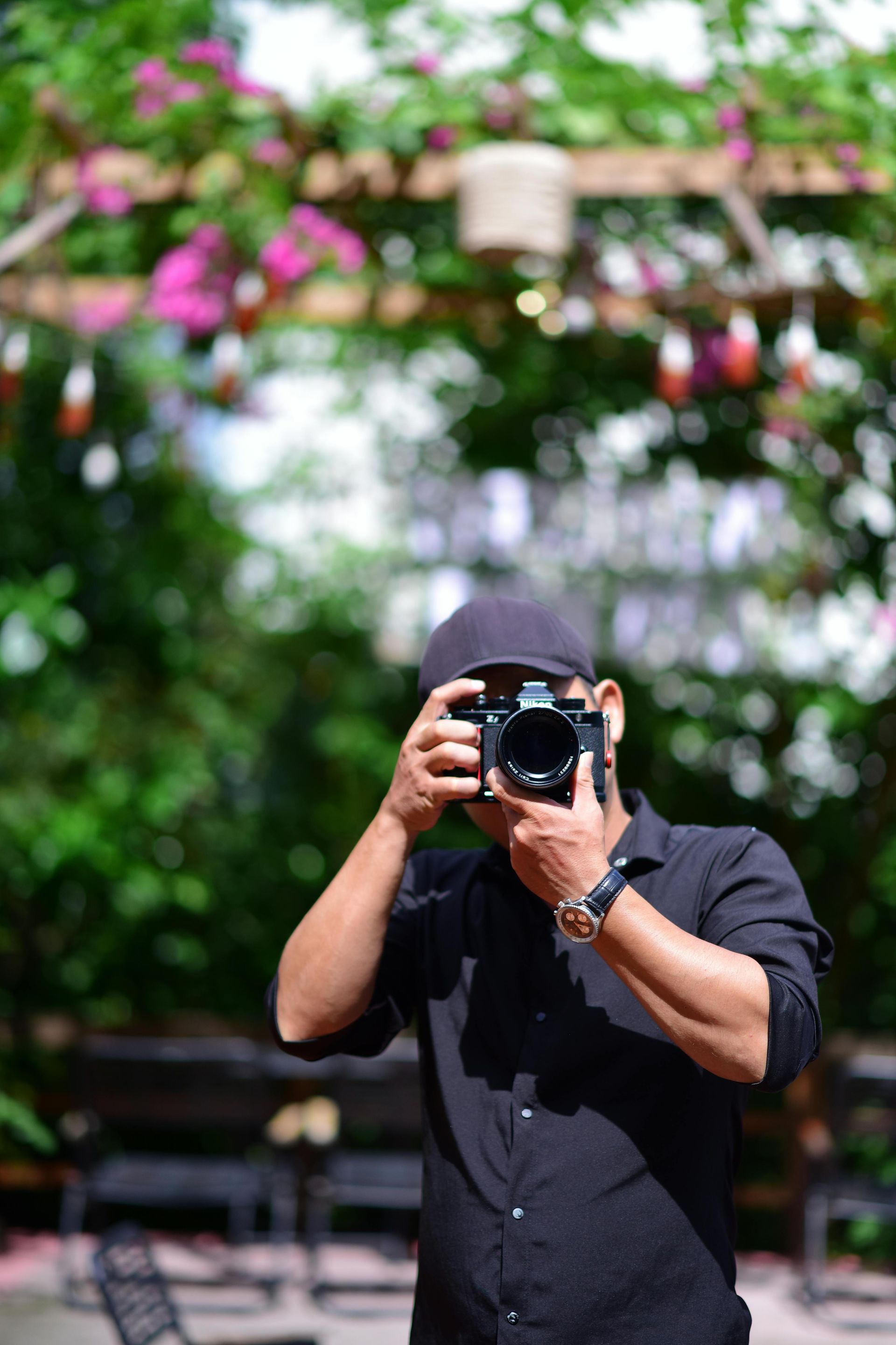 Photographer in black shirt and cap takes photo, outdoors with blurred greenery and flowers in background.