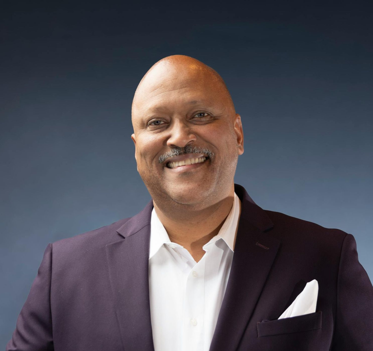 Man in a purple blazer and white shirt, smiling, with a blue background.