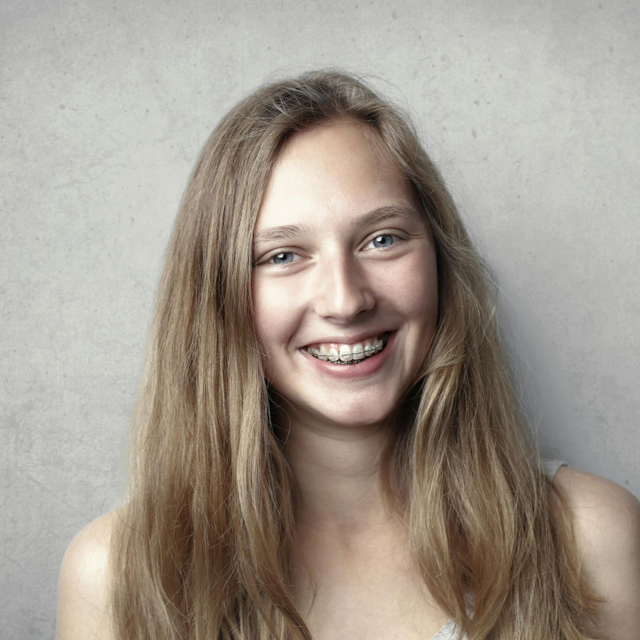 Woman with long, blonde hair and braces, smiling brightly, against a gray wall.