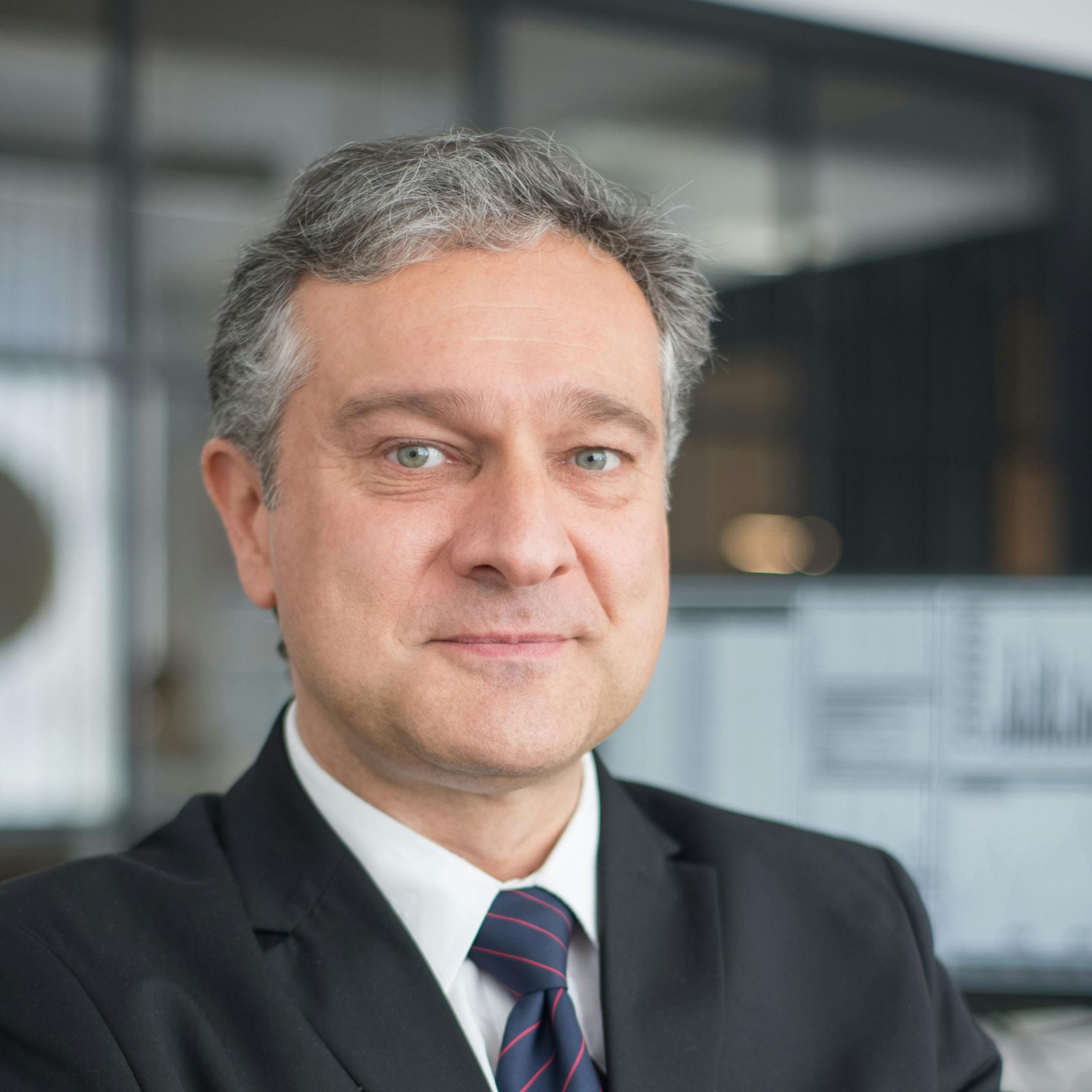 Man in suit smiles at the camera, indoors, blurred background.