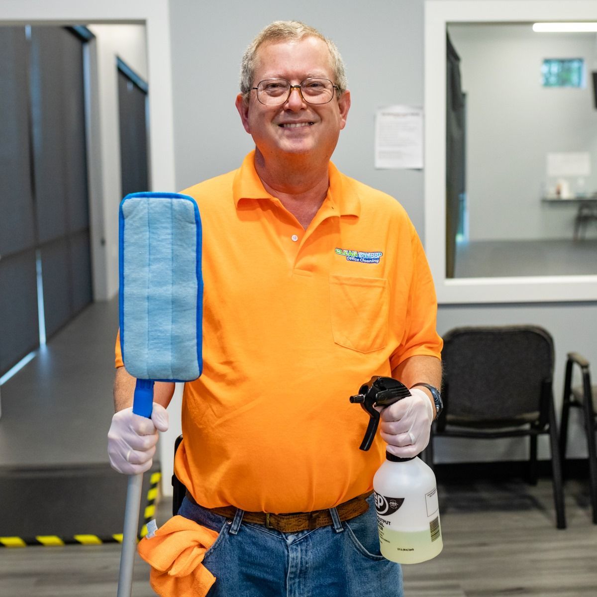 A man in an orange shirt is holding a mop and a spray bottle.
