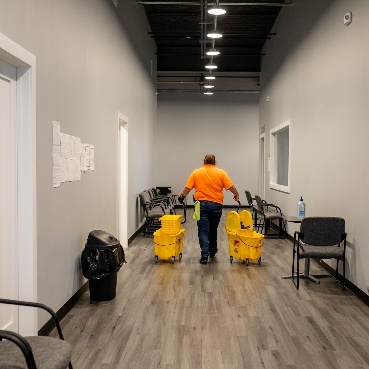 A man in an orange shirt is walking down a hallway carrying yellow buckets.