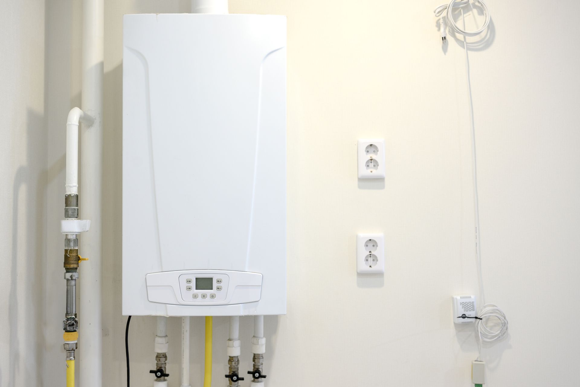 Wall-mounted white boiler with exposed pipes and outlets on a plain interior wall.