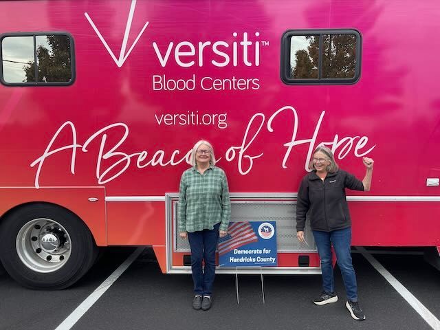 Two women stand by a Versiti Blood Centers mobile unit. The vehicle is pink with text,