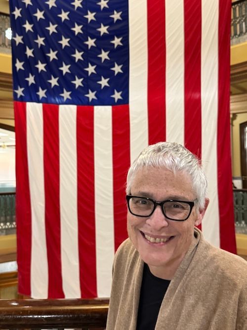 Woman with glasses smiles in front of a large American flag.