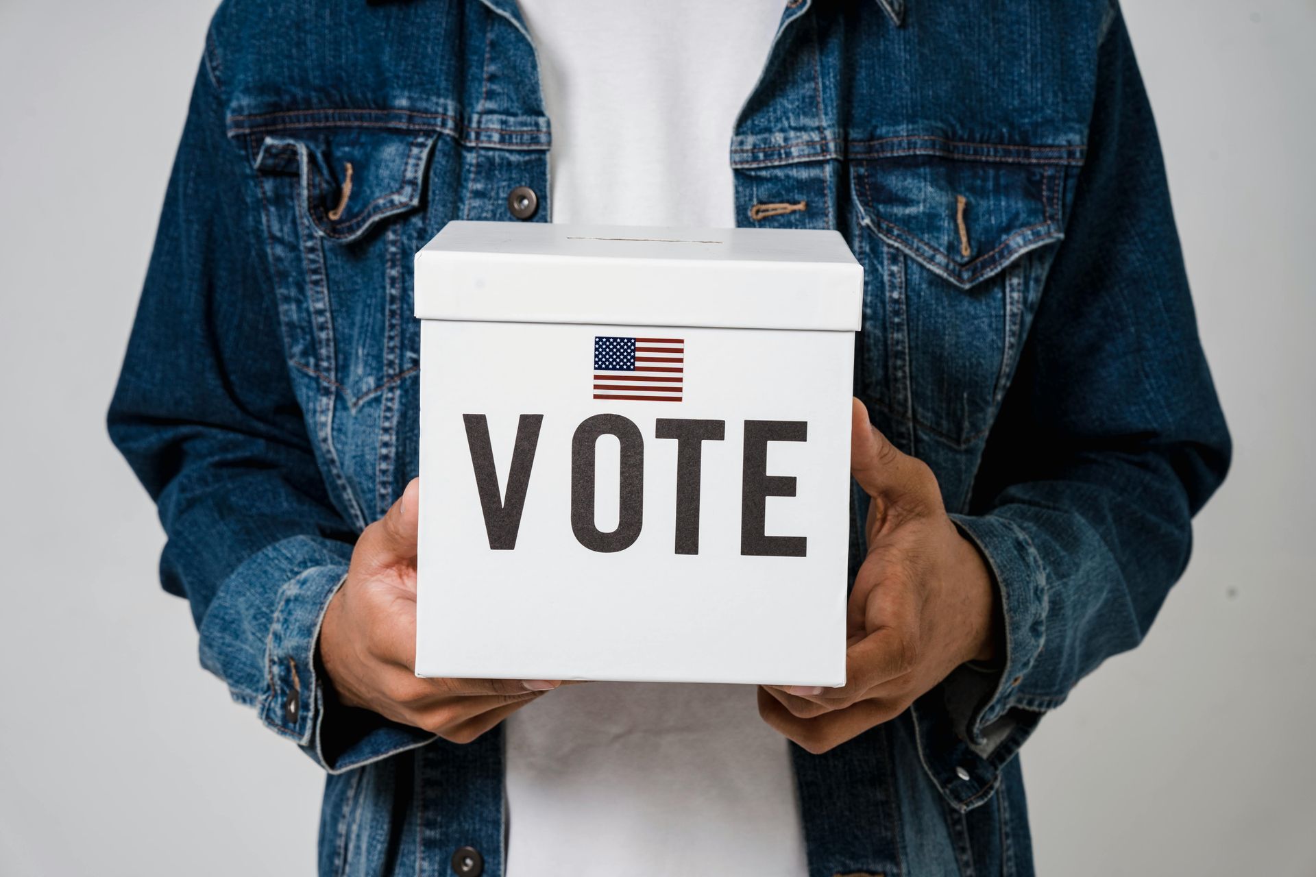 Person in denim jacket holding a white ballot box with “VOTE” and US flag printed on it.