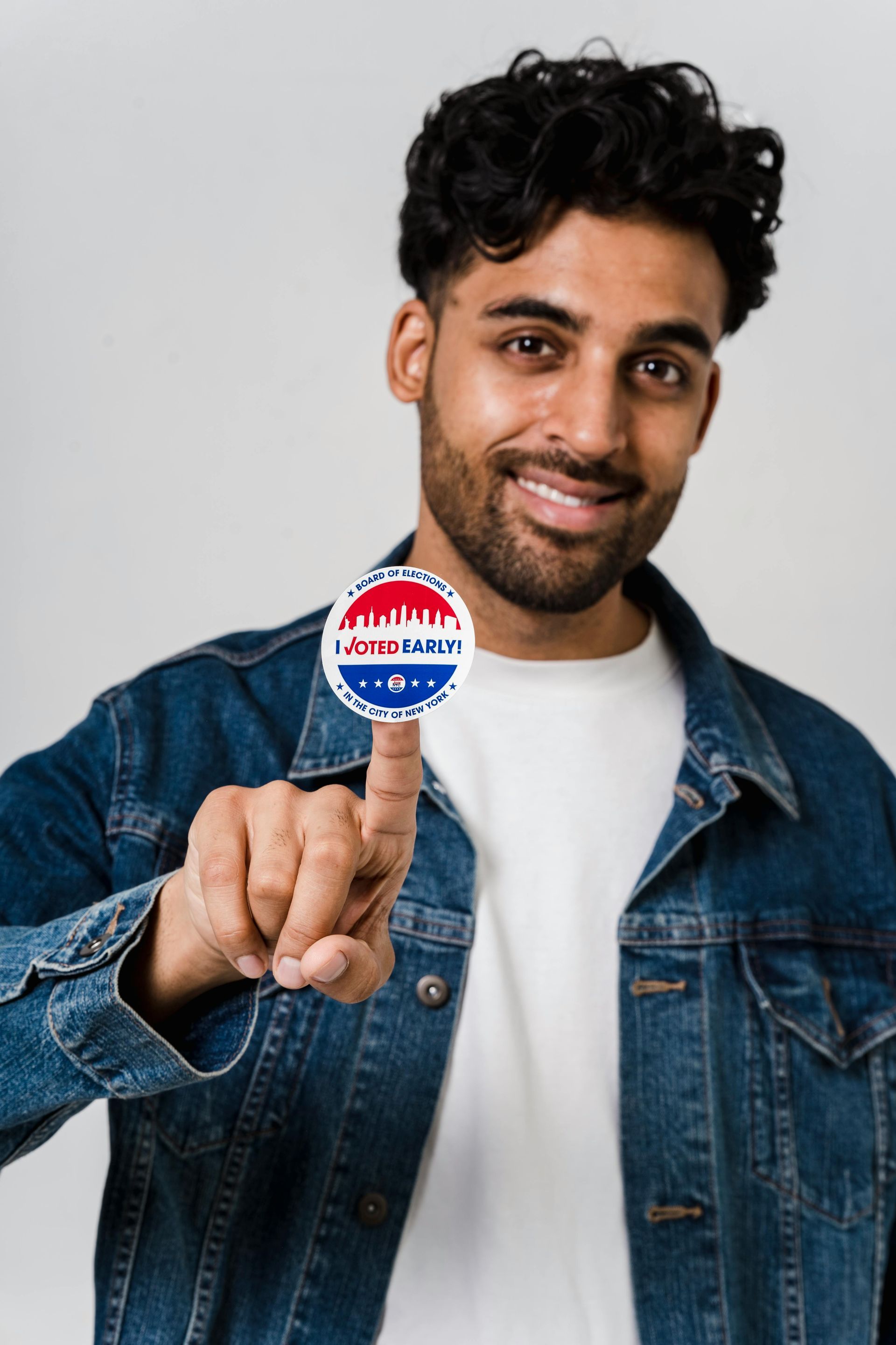 Man holds a New Jersey voting button, smiling. He's wearing a denim jacket.
