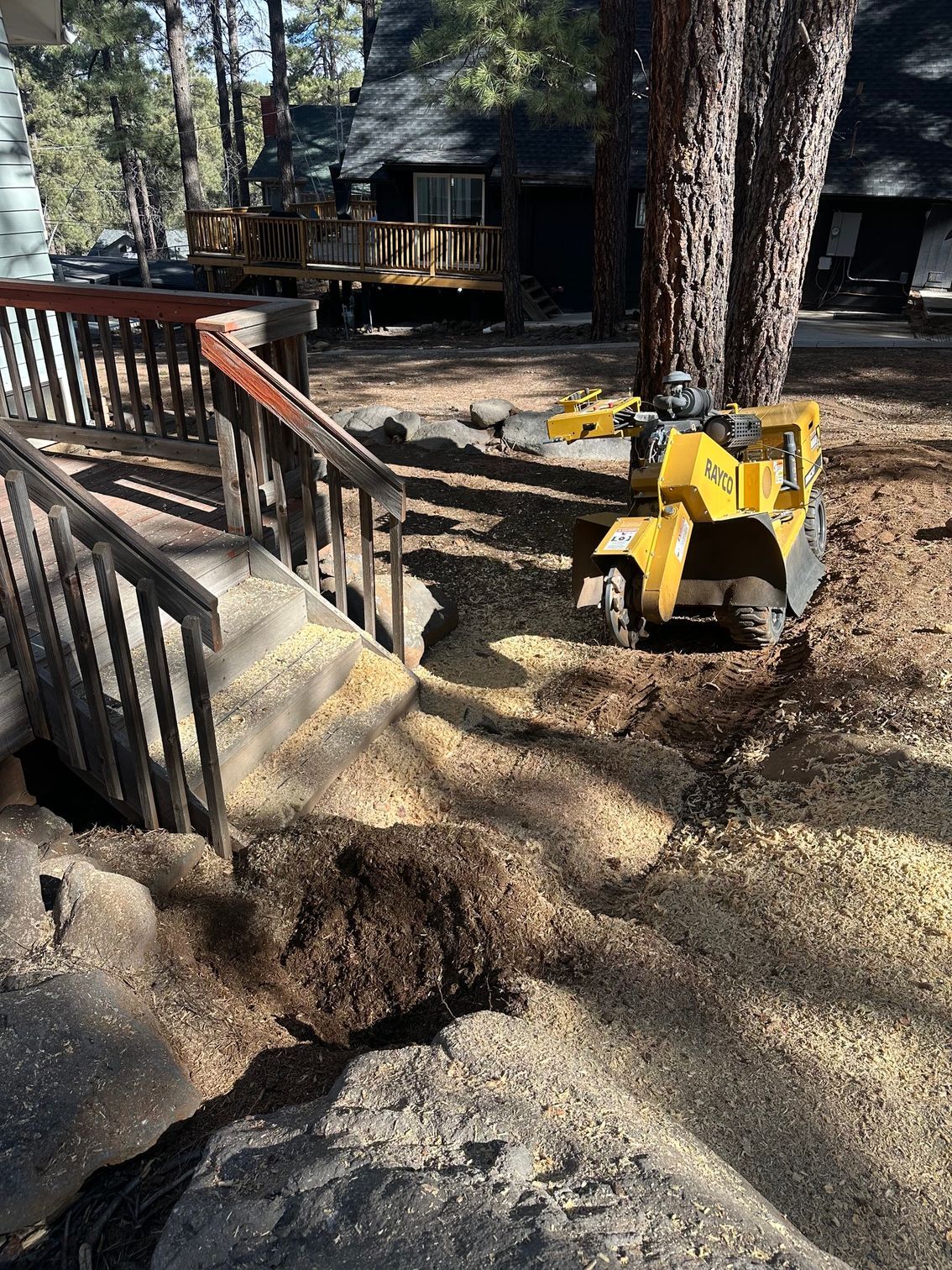 Yellow toy bulldozer on a dirt path beside a wooden deck and large trees in a wooded yard