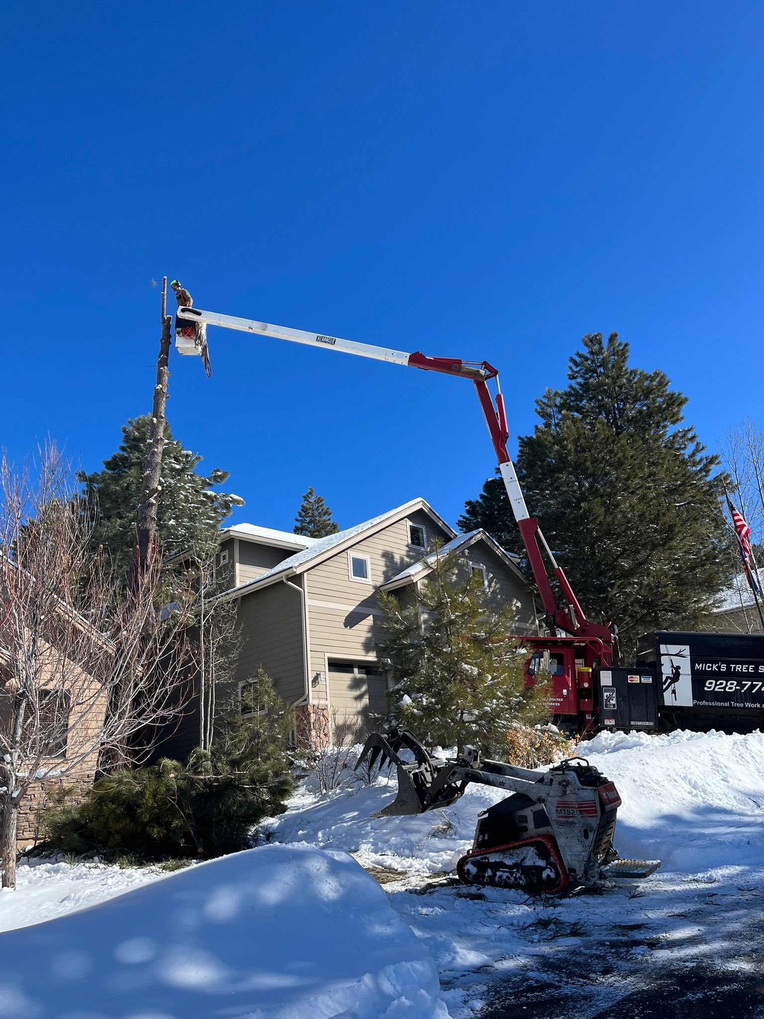 Worker in a cherry picker trimming a tree beside a house in a snowy yard under a clear blue sky