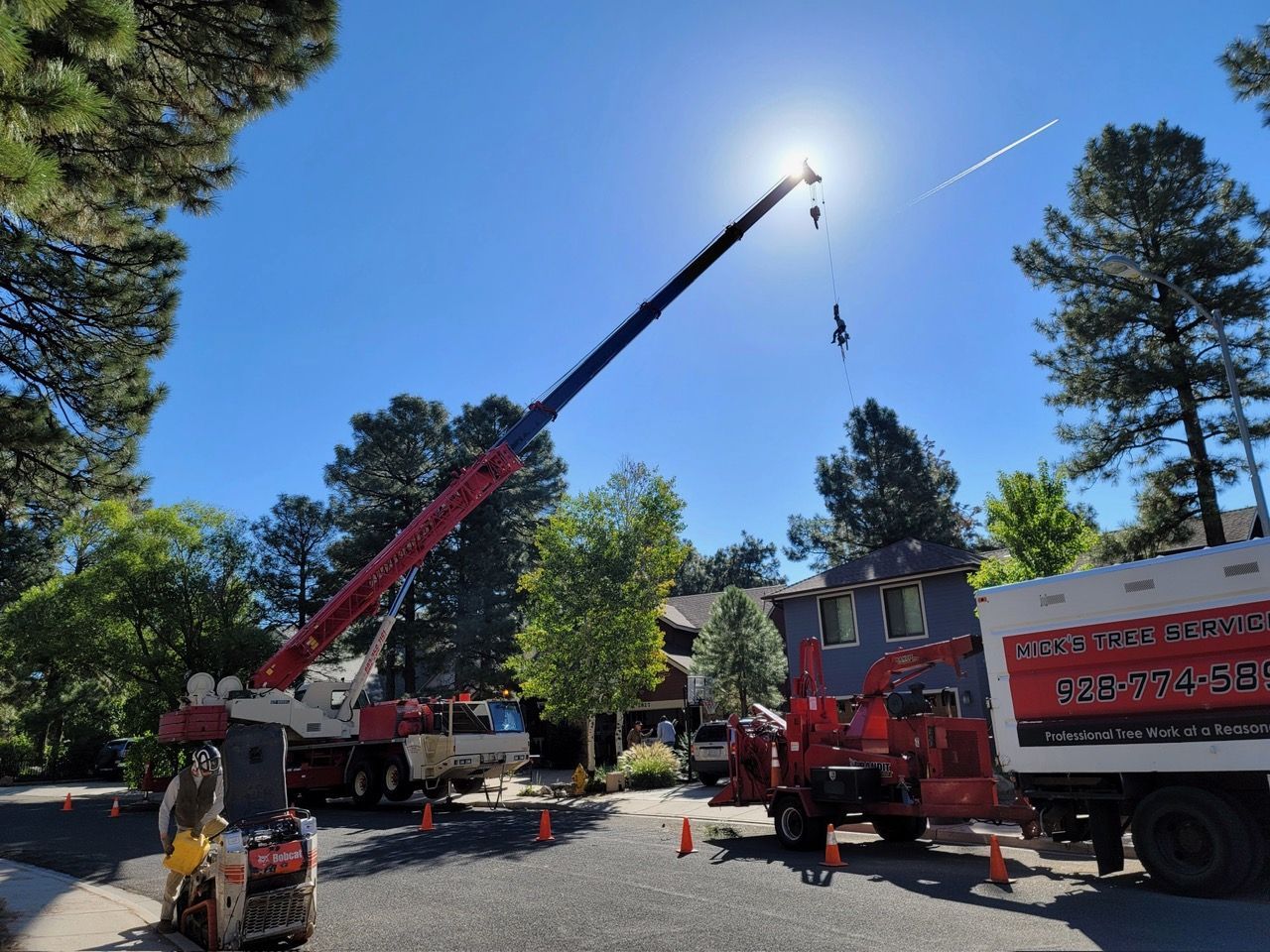 Crane lifting a bucket into a tree-lined street beside red emergency trucks on a sunny day