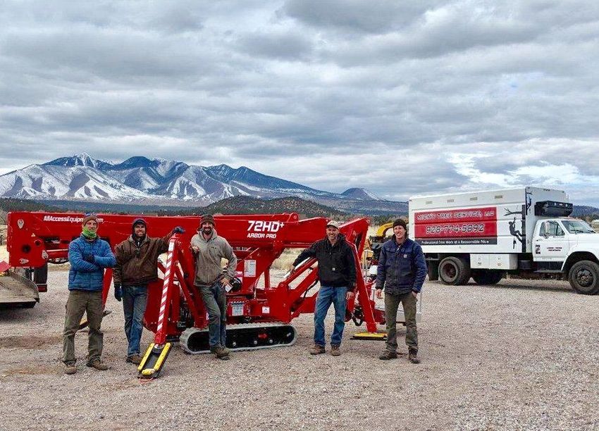 Group posing beside red machinery and a truck in a gravel lot with snowy mountains in the background