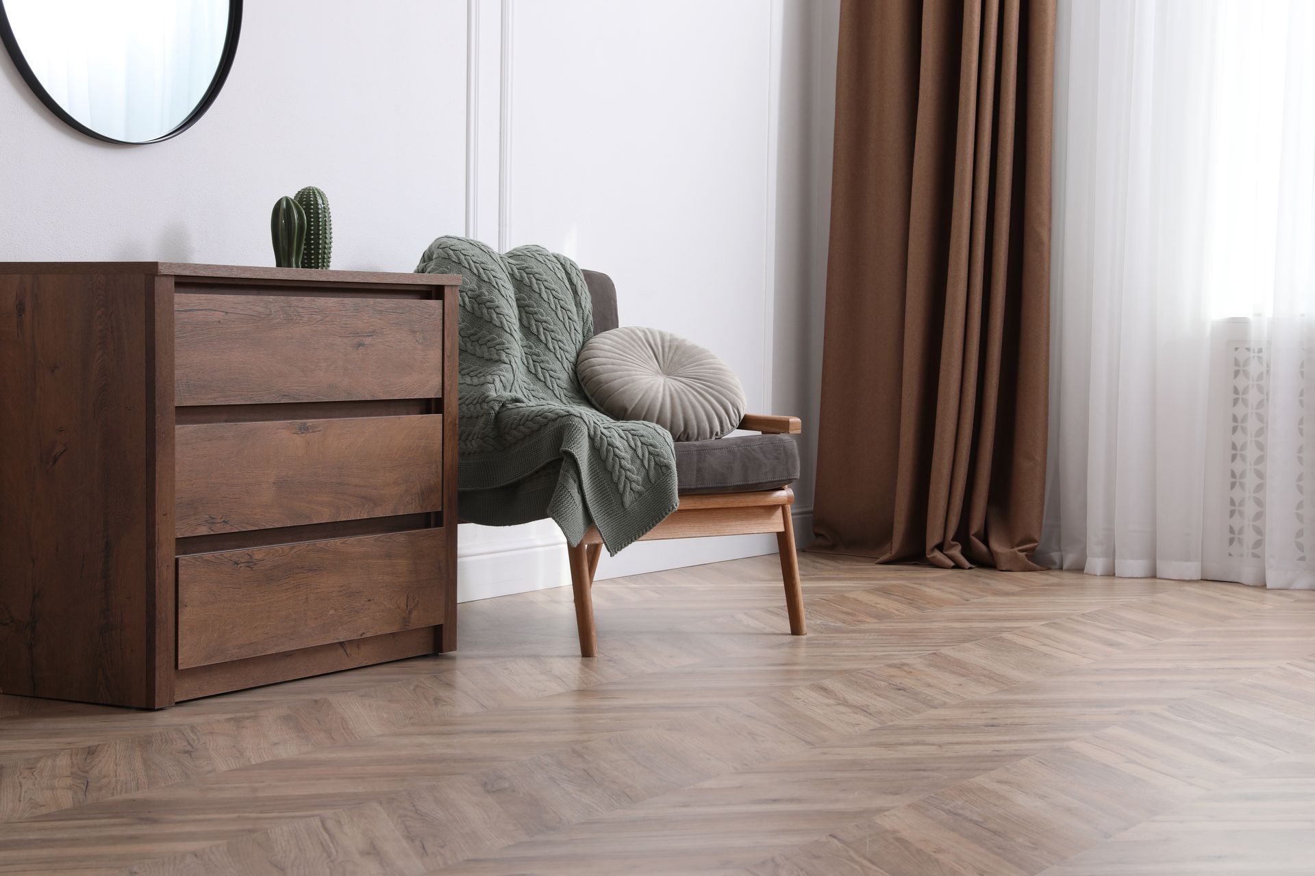 Wooden dresser, armchair with green blanket and pillow, near a window and curtains.