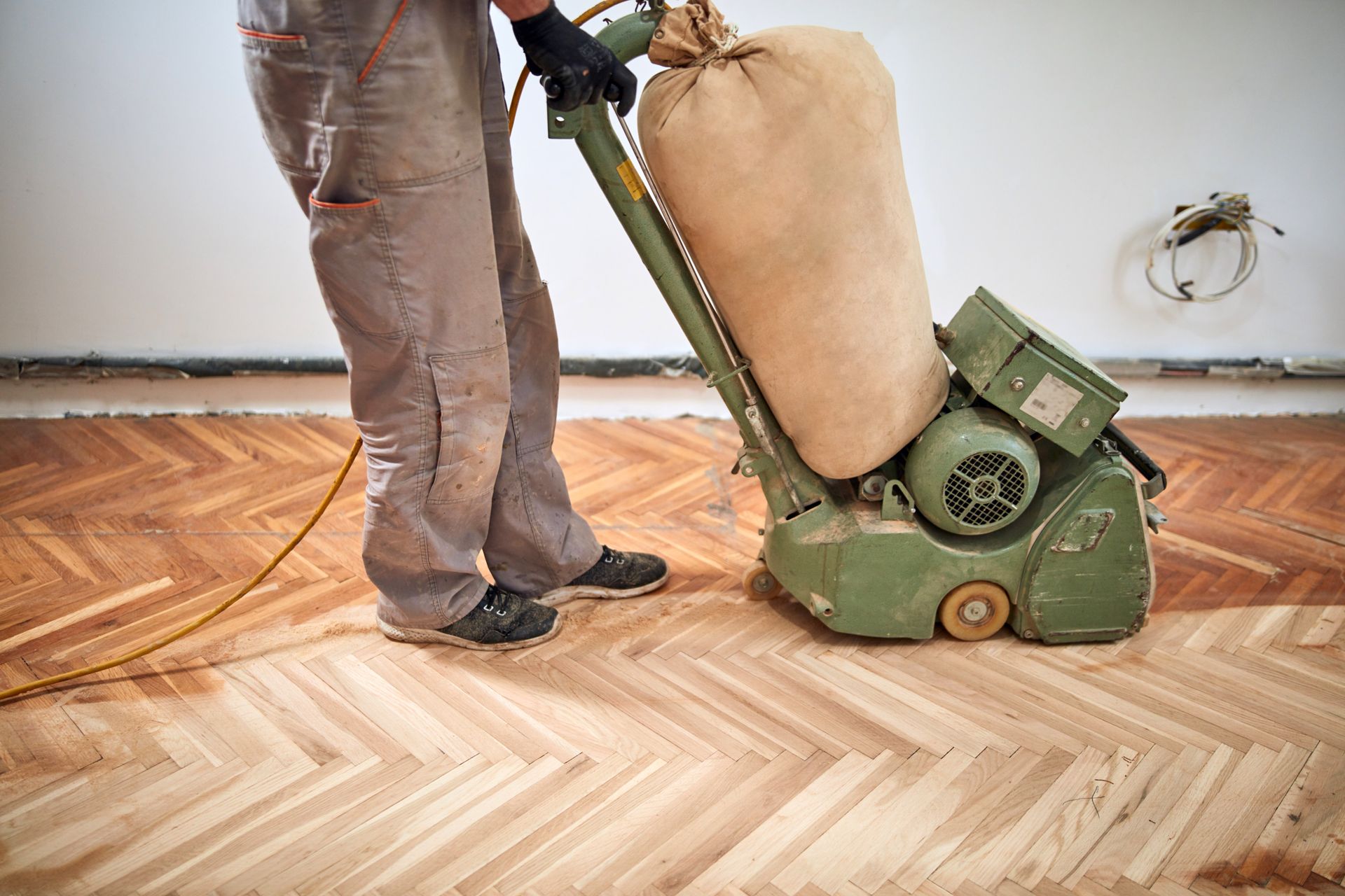 Person sanding a herringbone wood floor with a large sanding machine; tan bag collects sawdust.