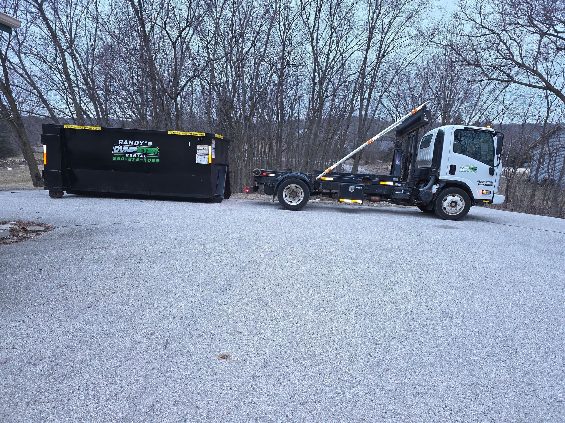 White truck with a black dumpster in front of a brick building on a sunny street.