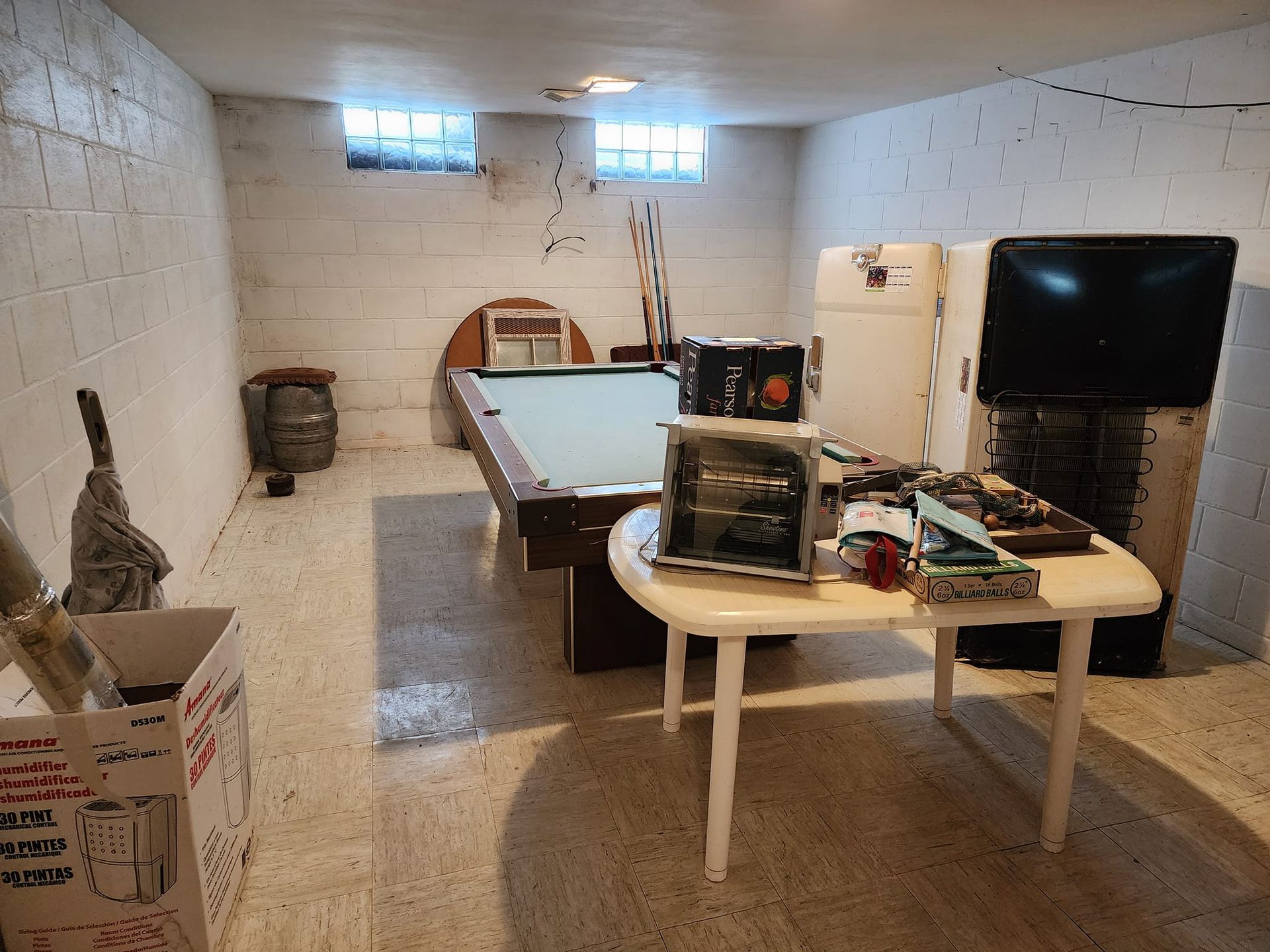 Basement with pool table, old appliances, and cluttered table. Concrete walls and small windows.