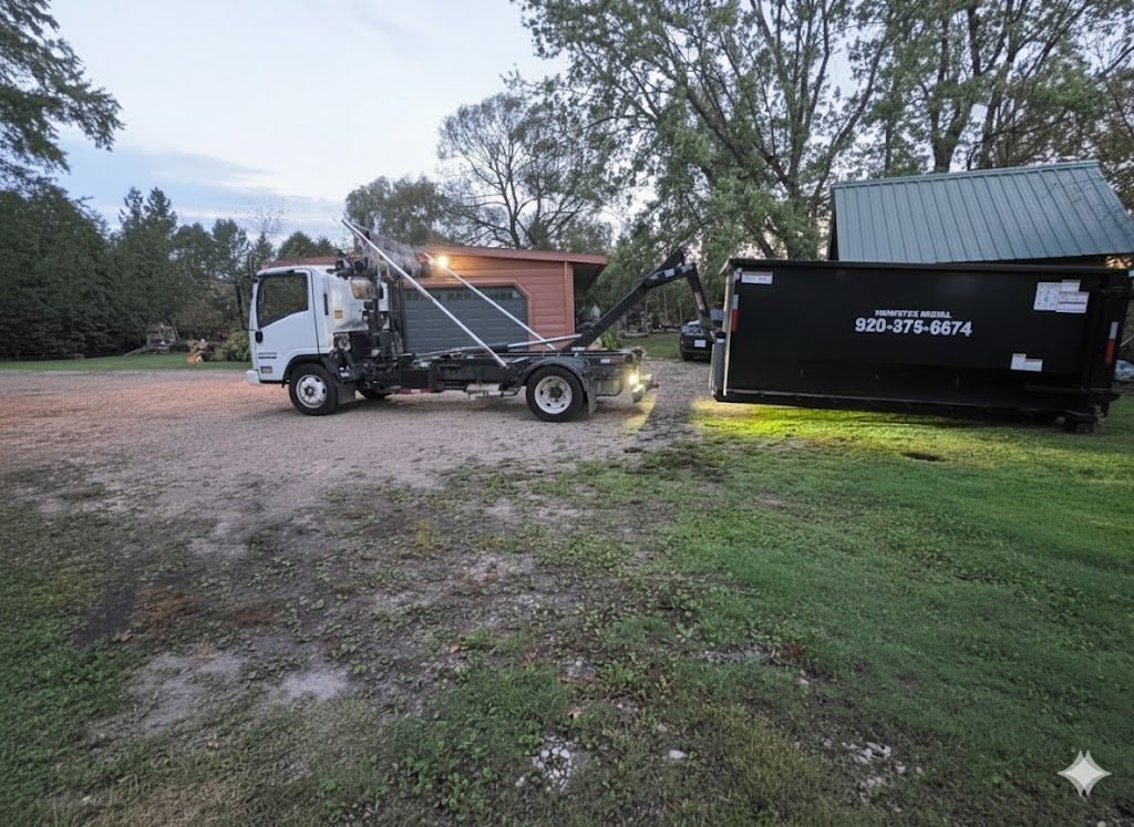 Truck lifting a black dumpster on a gravel driveway, next to green grass and a small building.