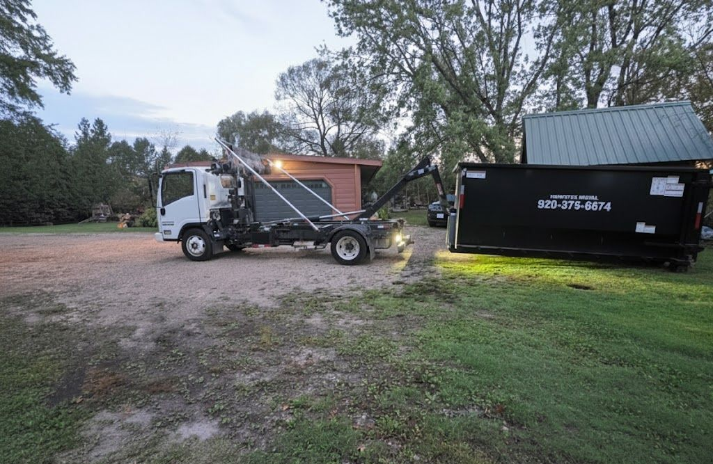 Truck with lift arm loading a black dumpster onto a gravel driveway beside a grassy yard, near dusk.