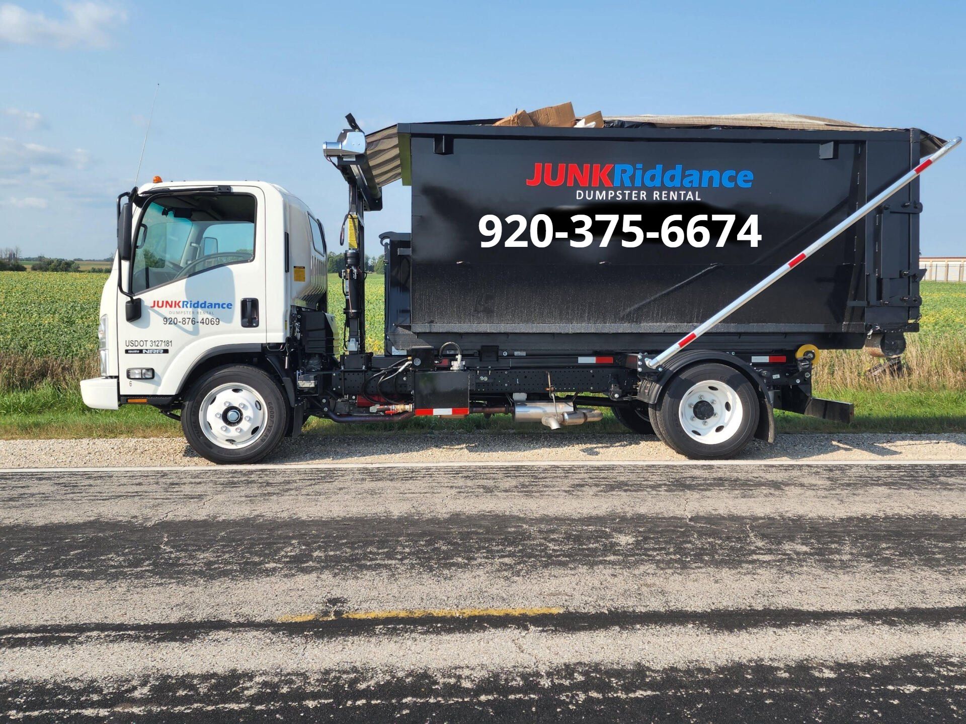 A white junk removal truck with a black container on a paved road. Contact number 920-375-6674 is visible.