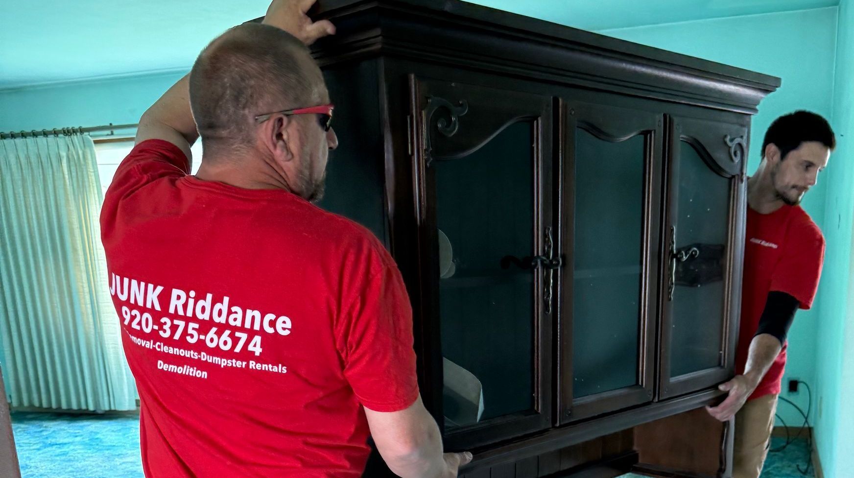 Two men in red shirts are carrying a large wooden cabinet removing junk in Sheboygan