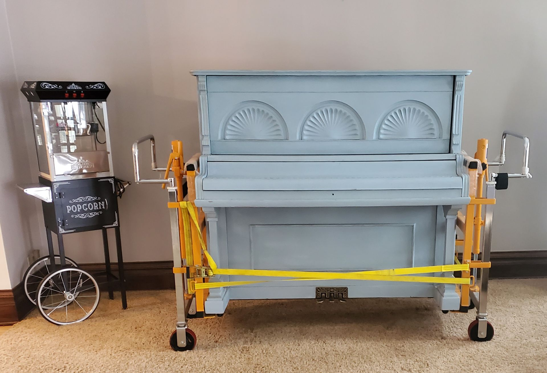 Popcorn machine and light blue piano with wheels against a beige wall. Yellow bands secure the piano.