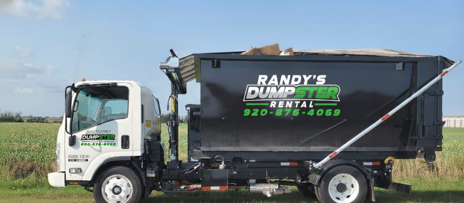 Truck with lift arm loading a black dumpster onto a gravel driveway beside a grassy yard, near dusk.