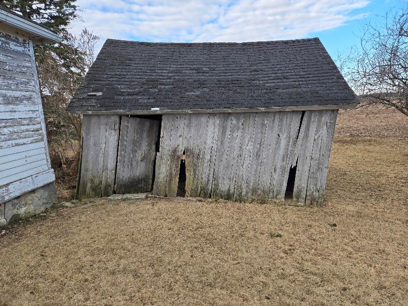 Weathered wooden shed with a slanted, gray roof and open doorway, in a field with brown grass.