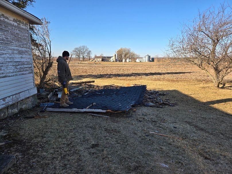 Person standing next to fallen roof debris near a weathered building and open field on a sunny day.