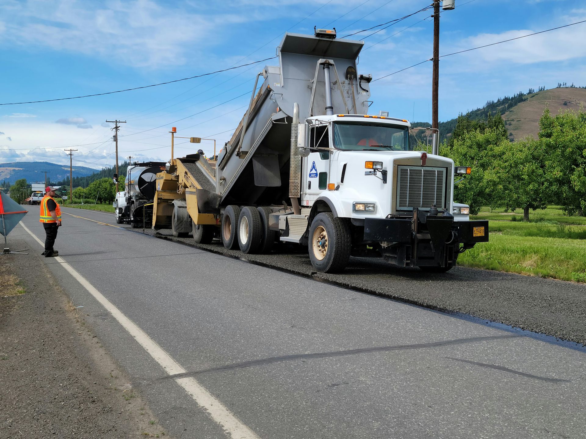 Road paving in progress. A dump truck pours gravel onto the road with worker in safety vest nearby. Hills in background.