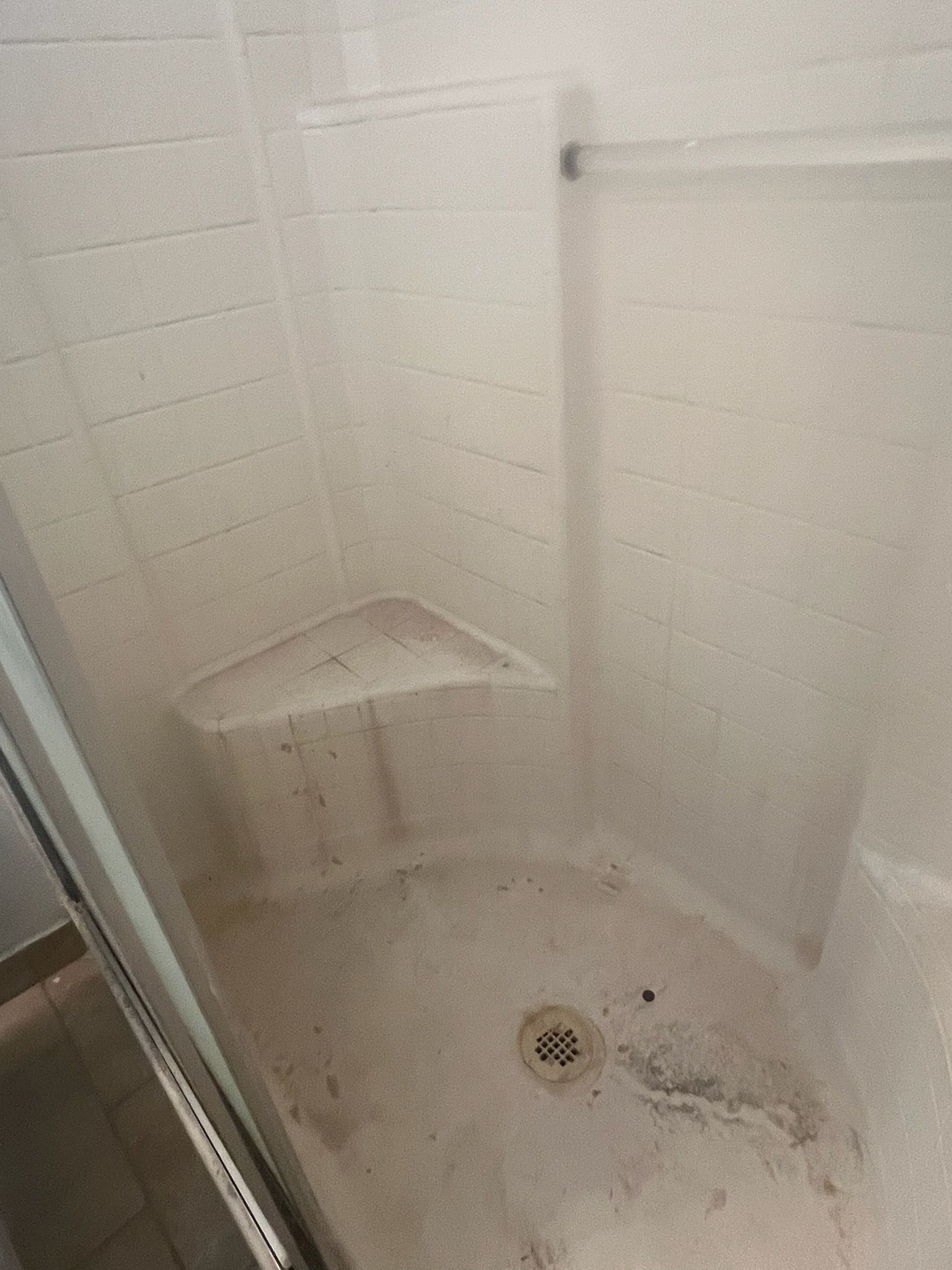 Shower interior, white walls, built-in corner shelf, drain in floor, signs of mildew.