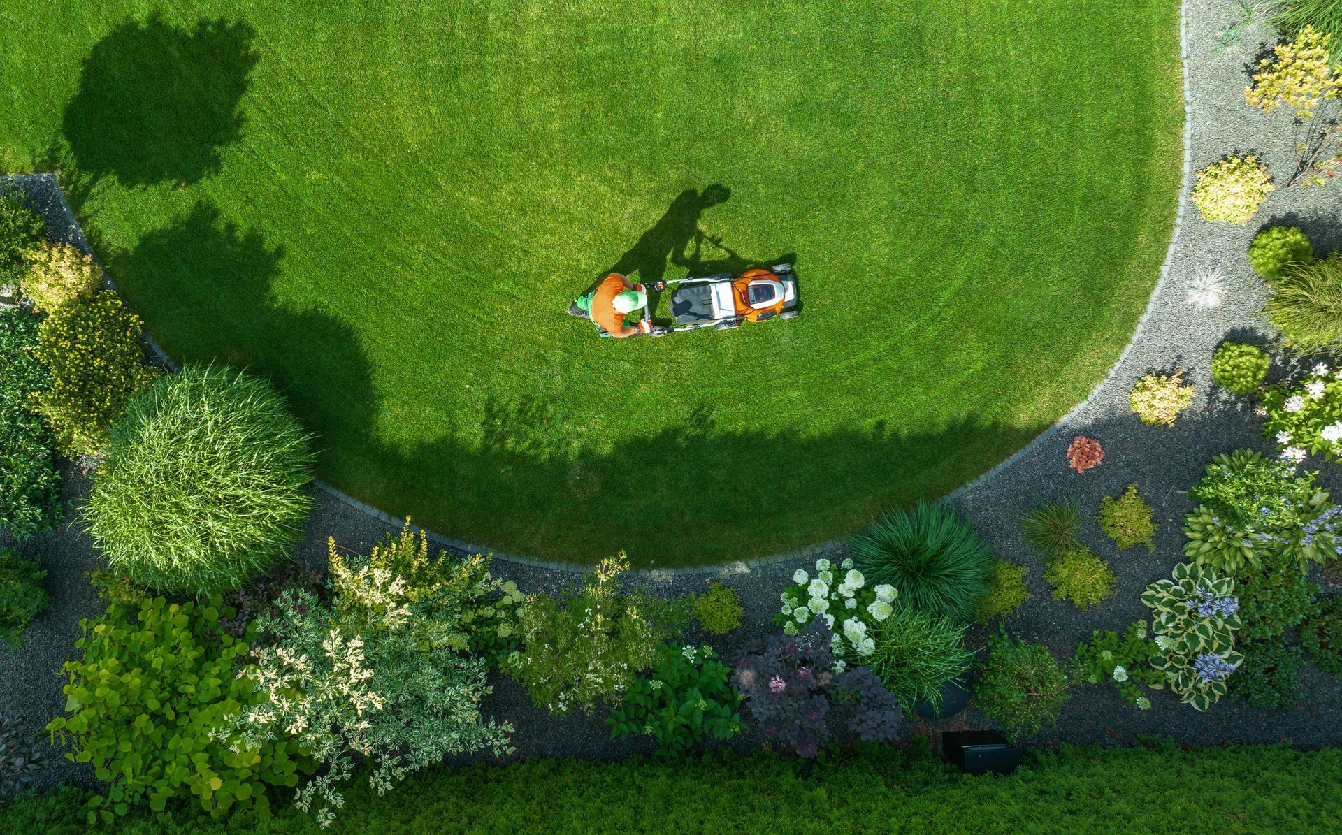 A gardener mows a well-maintained lawn, encircled by colorful plants and flowers in a residential yard.