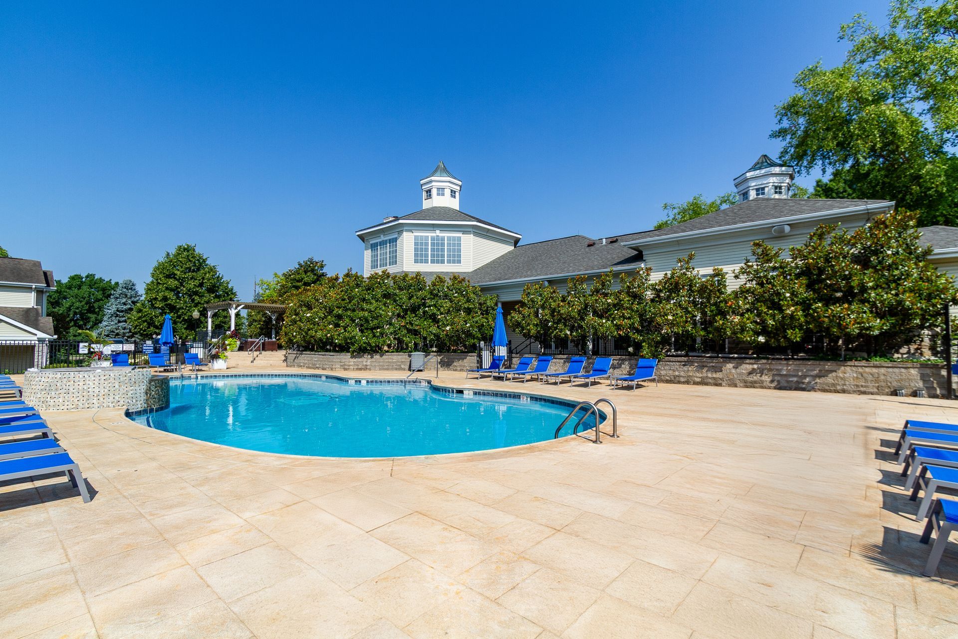 Swimming pool with blue water and lounge chairs, next to a building with a white exterior under a clear, blue sky. Explore scenic grounds and modern Apartments in Lexington, KY at Bridle Creek.