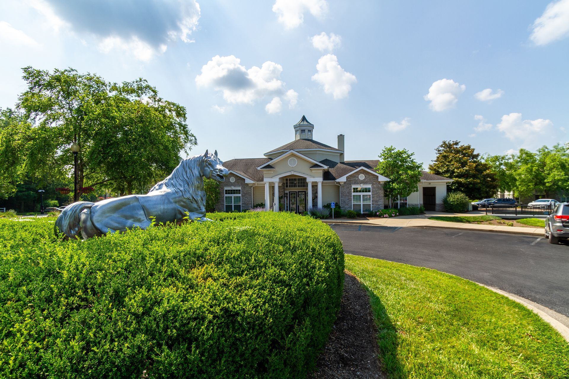 Exterior view of the resident clubhouse with a horse statue in front of a well-manicured green hedge and landscaping on a sunny day. Tour your future Lexington apartment rental at Bridle Creek today.