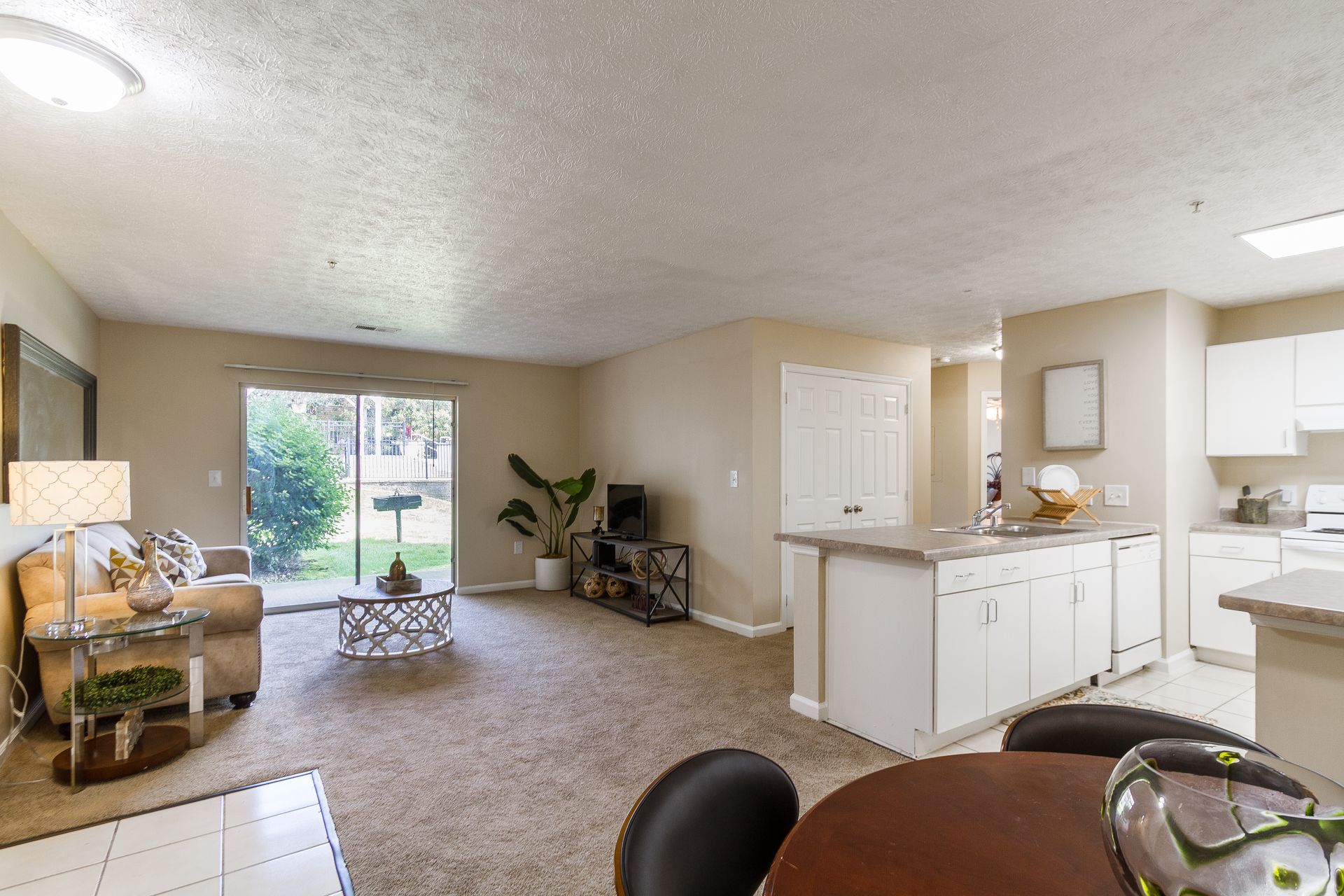 Spacious apartment living room with open kitchen, sliding glass door to yard, tan walls and carpet. Come explore inviting Apts in Lexington, KY at Bridle Creek Apartments.