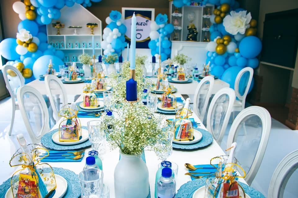 A long table with plates , glasses , and vases on it is decorated with blue and gold balloons.