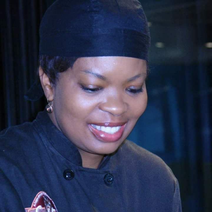 A woman in a chef 's uniform is smiling and wearing a black hat