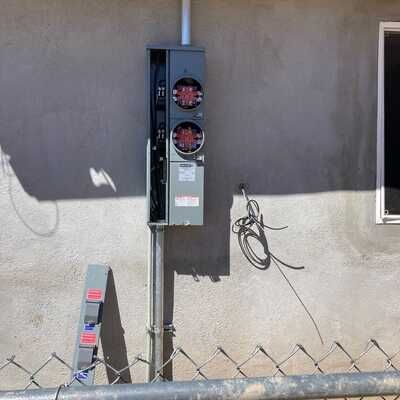 An electrical box is attached to the side of a building next to a chain link fence.