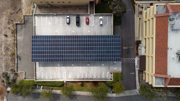Overhead view of a parking structure with solar panels and cars parked. Next to a beige building.