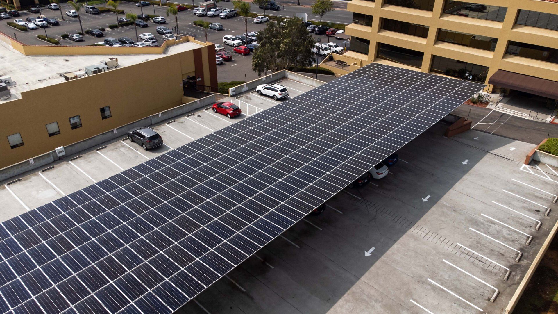 Solar panels cover a parking lot. Cars are parked beneath the panels, near a building.