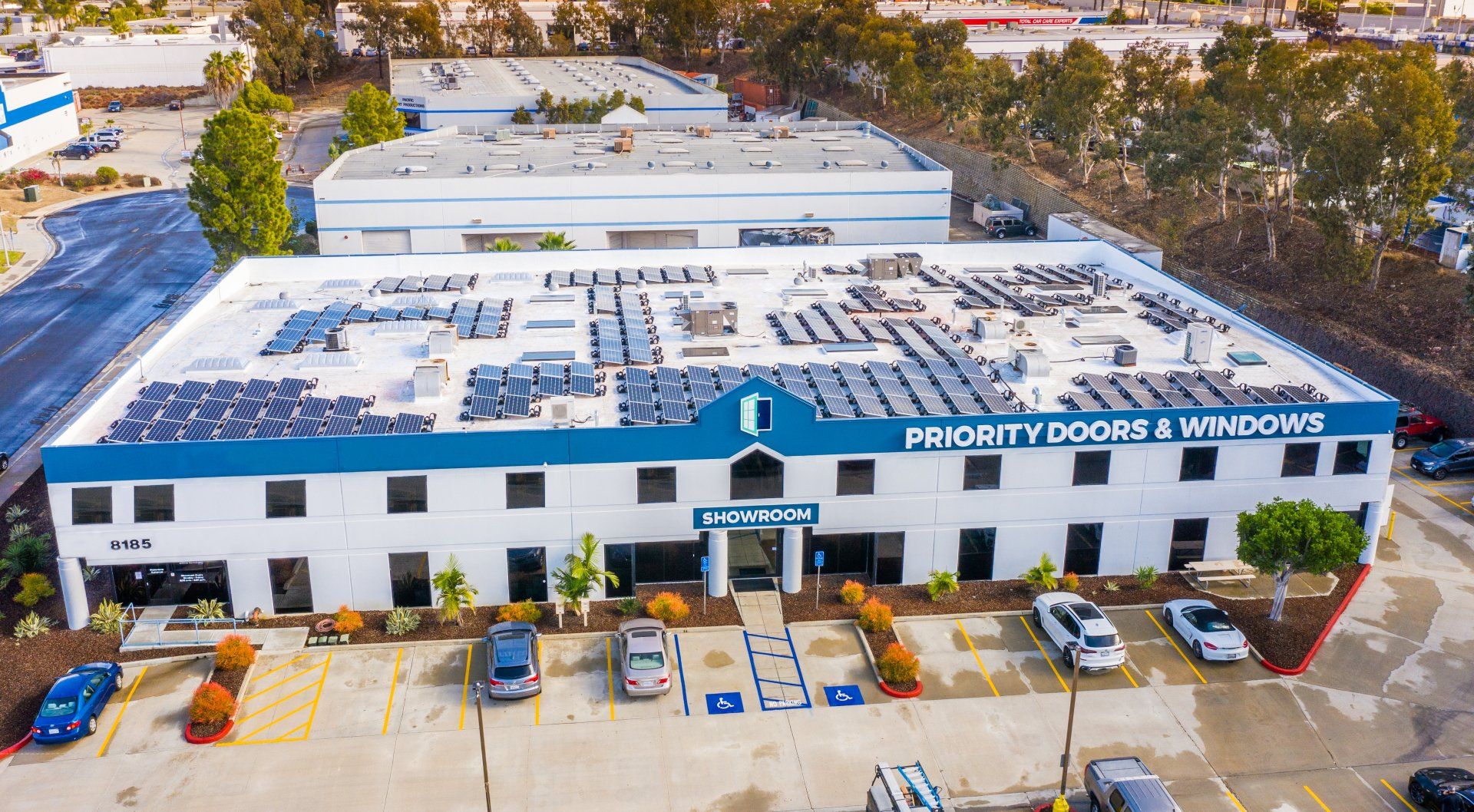 Exterior of Priority Doors & Windows building with solar panels on roof, blue and white facade, parked cars.