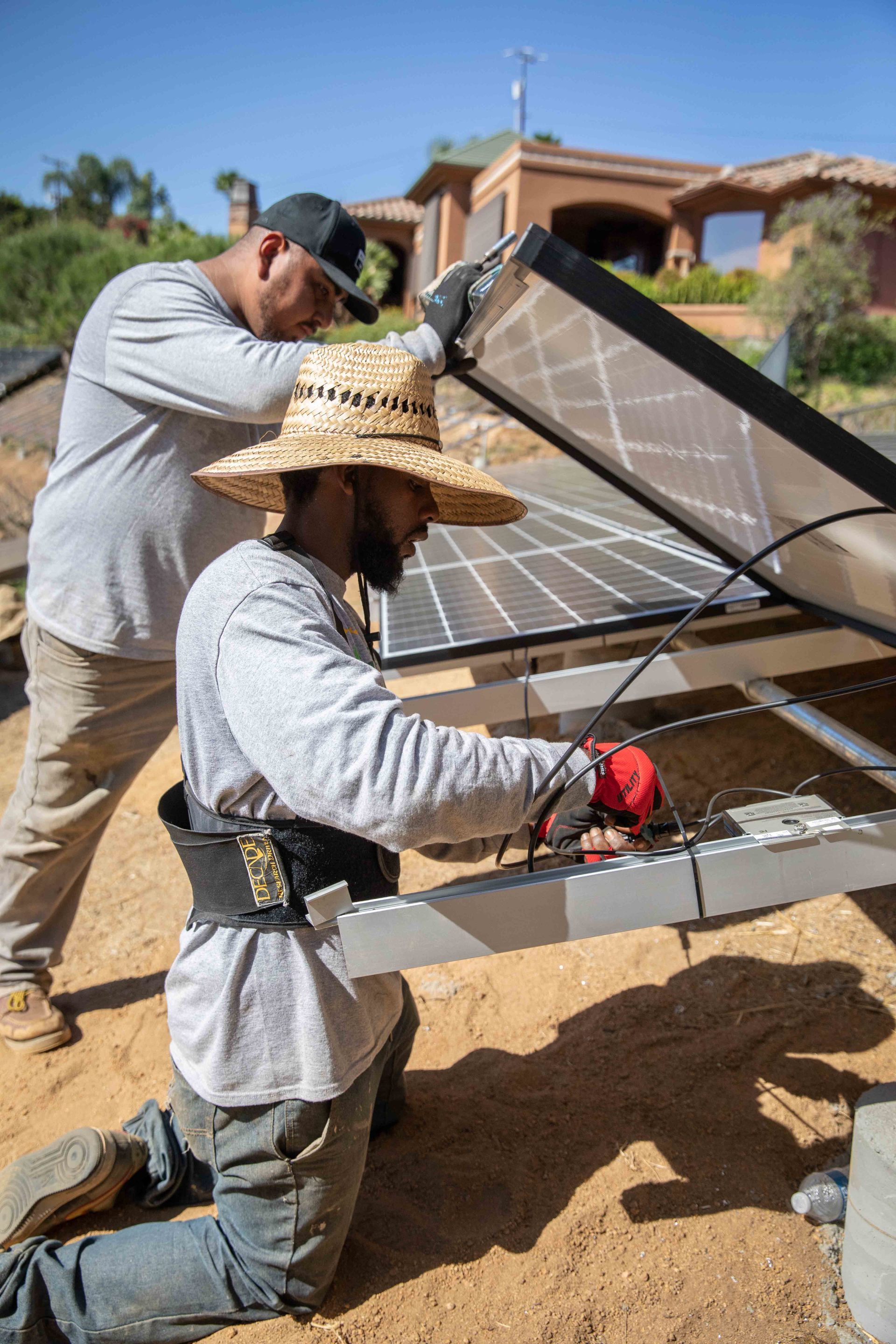 Two men installing solar panels on a rooftop. One kneels, connecting wires, other holds a panel, sunny outdoor setting.