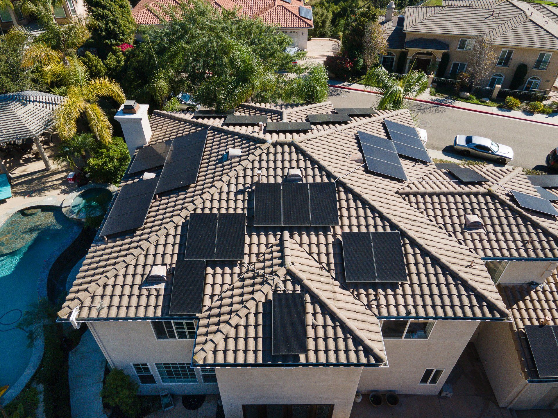 Solar panels on a tan-tiled roof of a large house, with a pool and other houses visible.