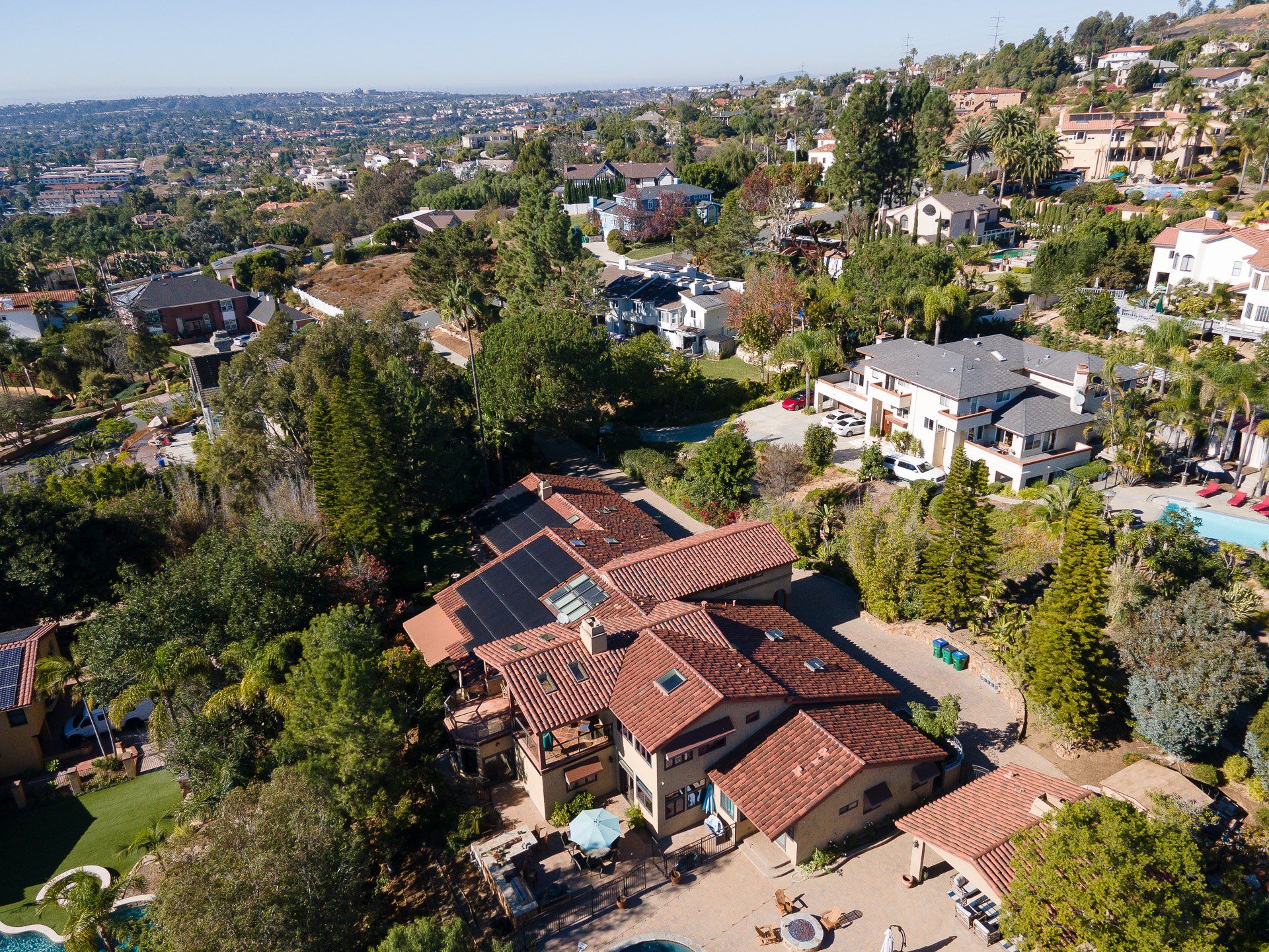 Aerial view of large, multi-roofed house with solar panels, overlooking a hillside neighborhood.