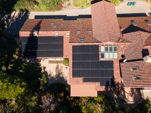 Overhead view of a house with solar panels on a clay tile roof, surrounded by trees.