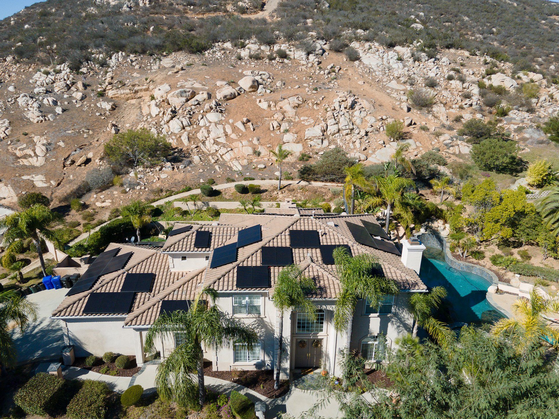 House with solar panels on the roof, palm trees, and a pool against a rocky hillside.