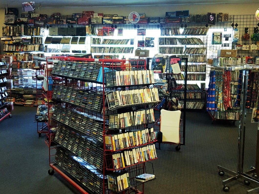 Inside a video rental store, movies line shelves and racks; a few customers.