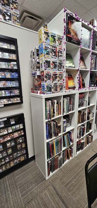 A store with bookshelves, Funko Pop figures, and a display case of cards. Carpeted floors.