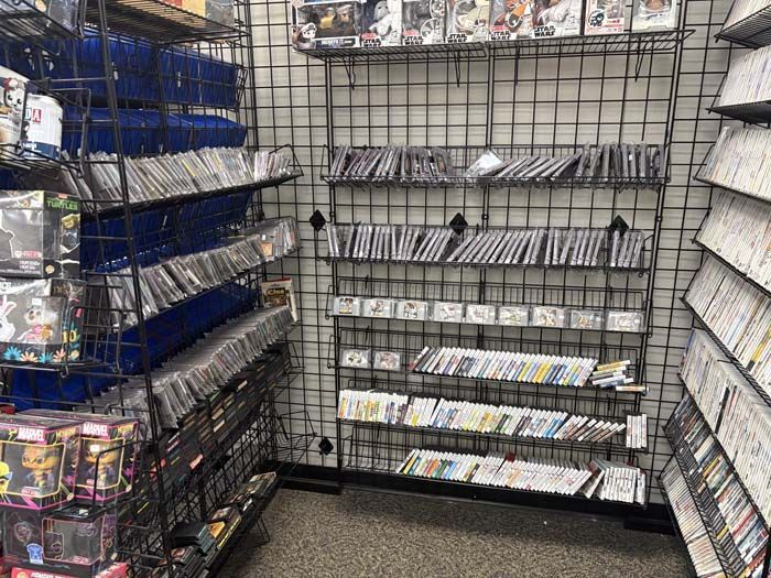 Shelves filled with video games inside a store; black metal shelving, blue baskets, beige floor.
