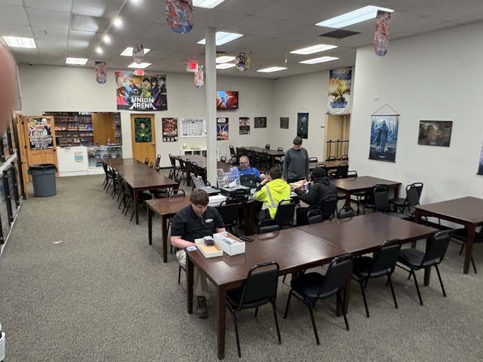 Game store interior with long tables, people playing, and fantasy/comic posters.