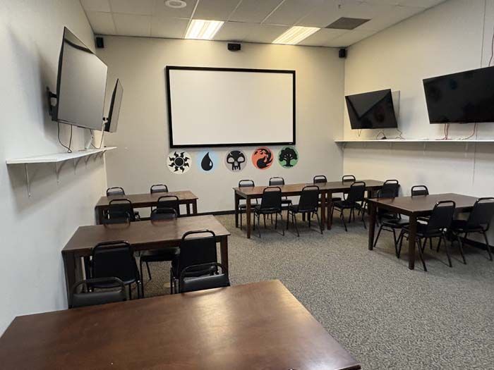 Classroom with tables, chairs, TVs, projector screen, and game-related symbols on wall.