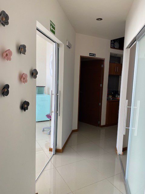 Pasillo con puertas abiertas, con flores en la pared, señal de salida de emergencia y suelo de baldosas.